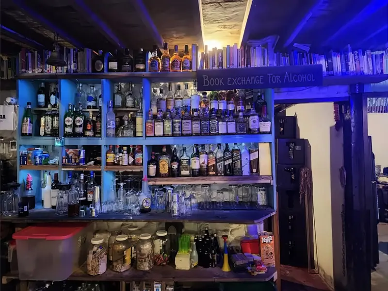 La estantería del bar iluminada en azul, con botellas, libros y el letrero «Book exchange for alcohol».