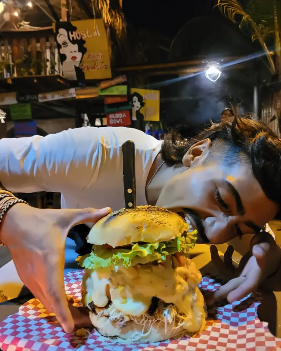 Un huésped se acerca a una hamburguesa enorme con queso derretido, sobre un mantel de cuadros rojos y blancos.