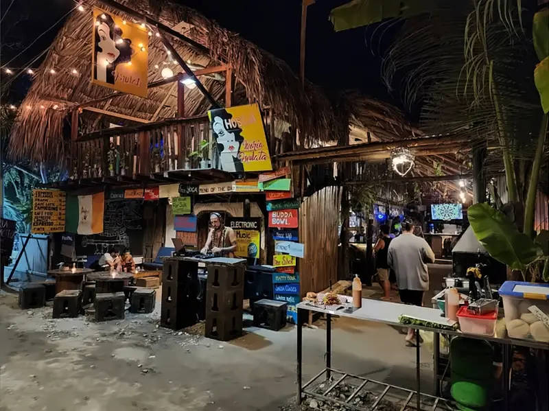El bar de noche: un DJ bajo el balcón de palma, banquitos de caja y letreros como «Budda Bowl» y «Electronic Music».