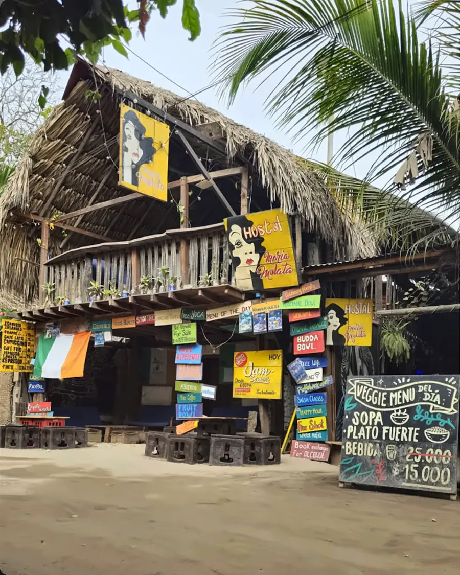 Fachada de María Mulata de día: techo de palma, balcón de madera, el letrero amarillo «Hostal María Mulata» y decenas de letreros pintados a mano sobre la calle de arena.