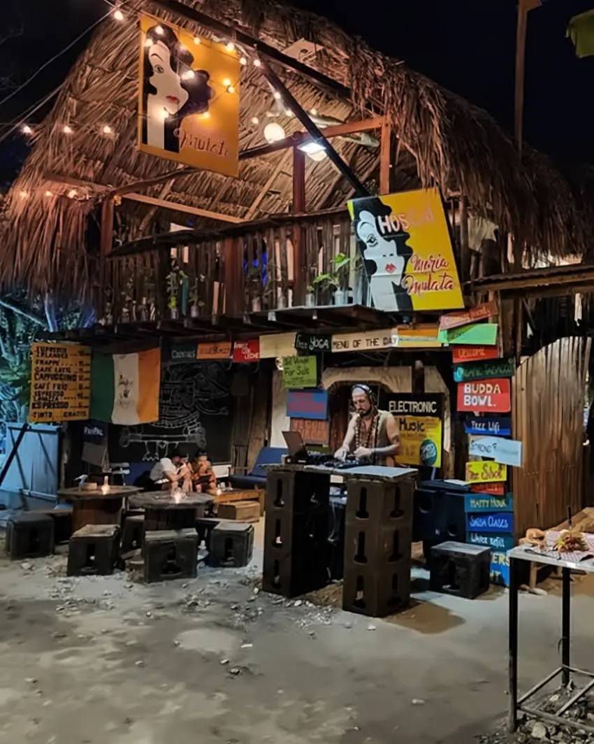 La misma fachada de noche: luces encendidas, letreros iluminados y un DJ tocando bajo el balcón de palma.