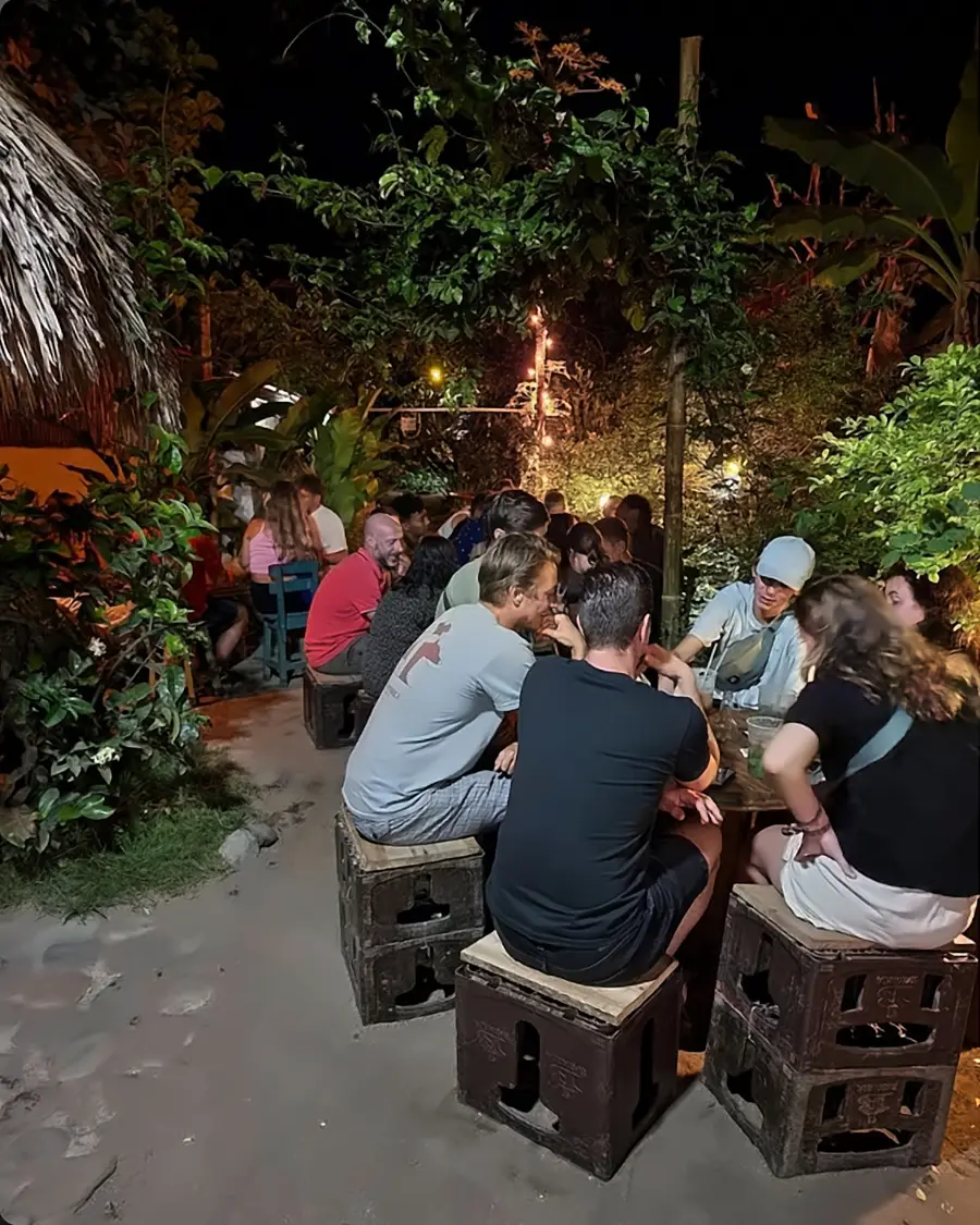 Viajeros conversando en banquitos de caja en el jardín tropical iluminado de noche.