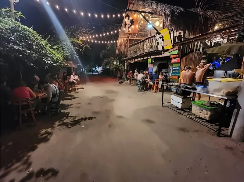La calle de arena frente al hostal de noche, con guirnaldas de luces y gente comiendo en mesas a ambos lados.