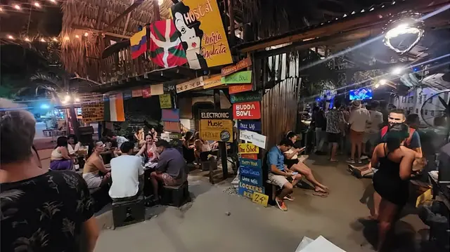 Panorámica de una noche llena: gente en banquitos, la pared de letreros y el bar encendido.
