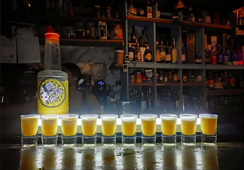 Una fila de diez shots amarillos frente a una botella de María Mulata y la estantería del bar.