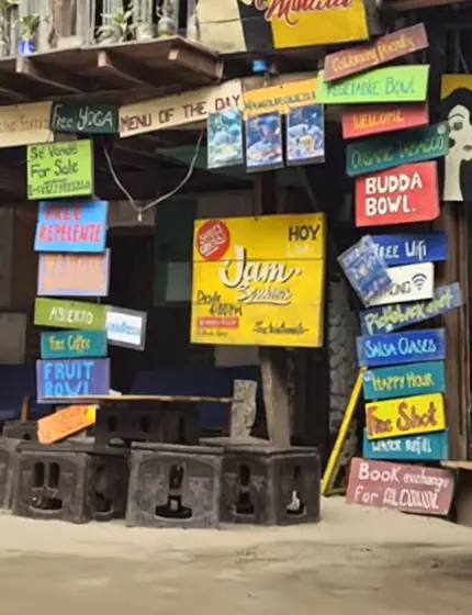 Detalle de la pared de letreros: el cartel amarillo de «Jam Sessions» y la columna de tablas pintadas —«Budda Bowl», «Free WiFi», «Salsa clases», «Happy hour», «Free shot», «Water refill», «Book exchange for alcohol»— junto al letrero «Hostal María Mulata».